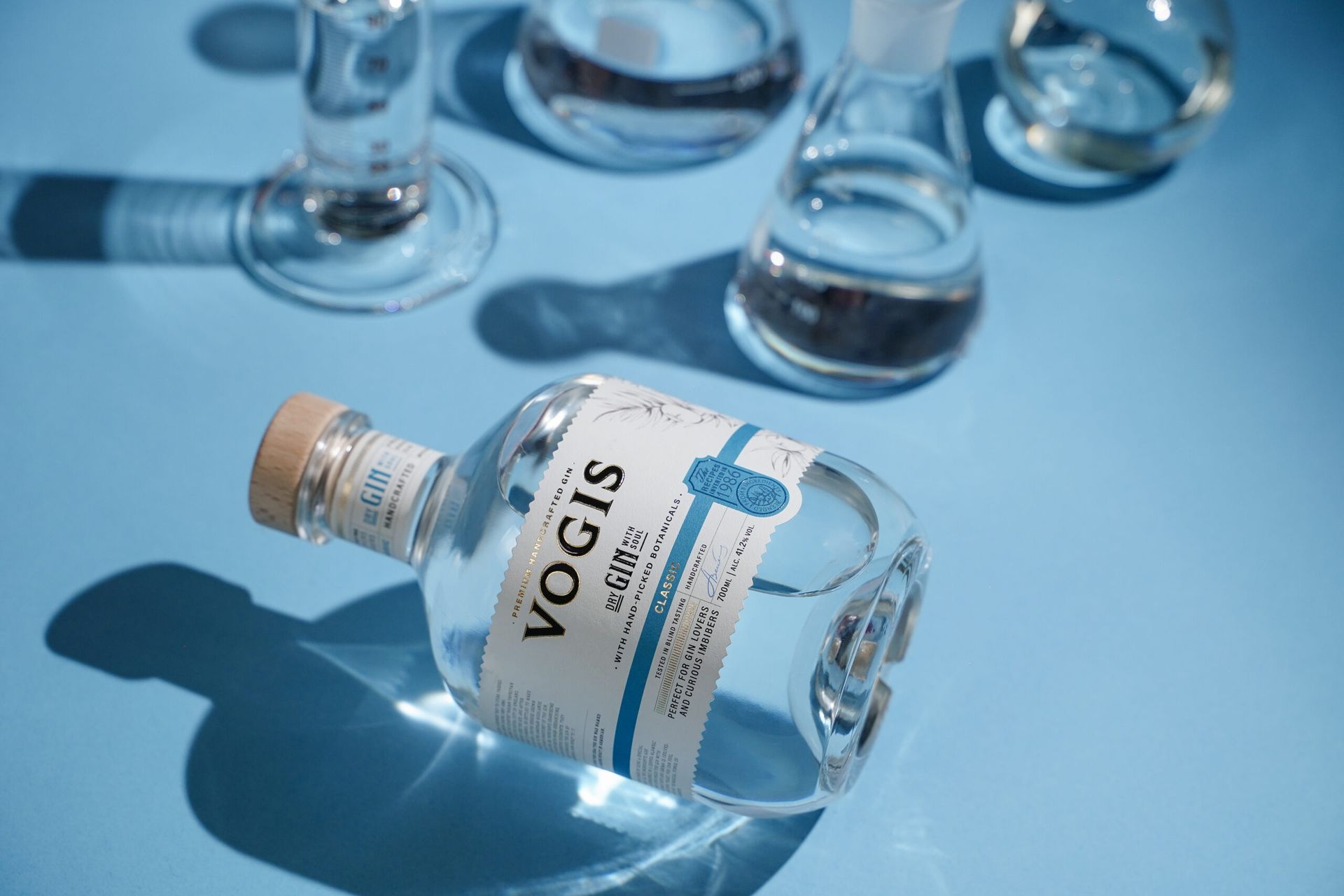 Vogis Classic Dry Gin bottle lying among glassware on a blue background